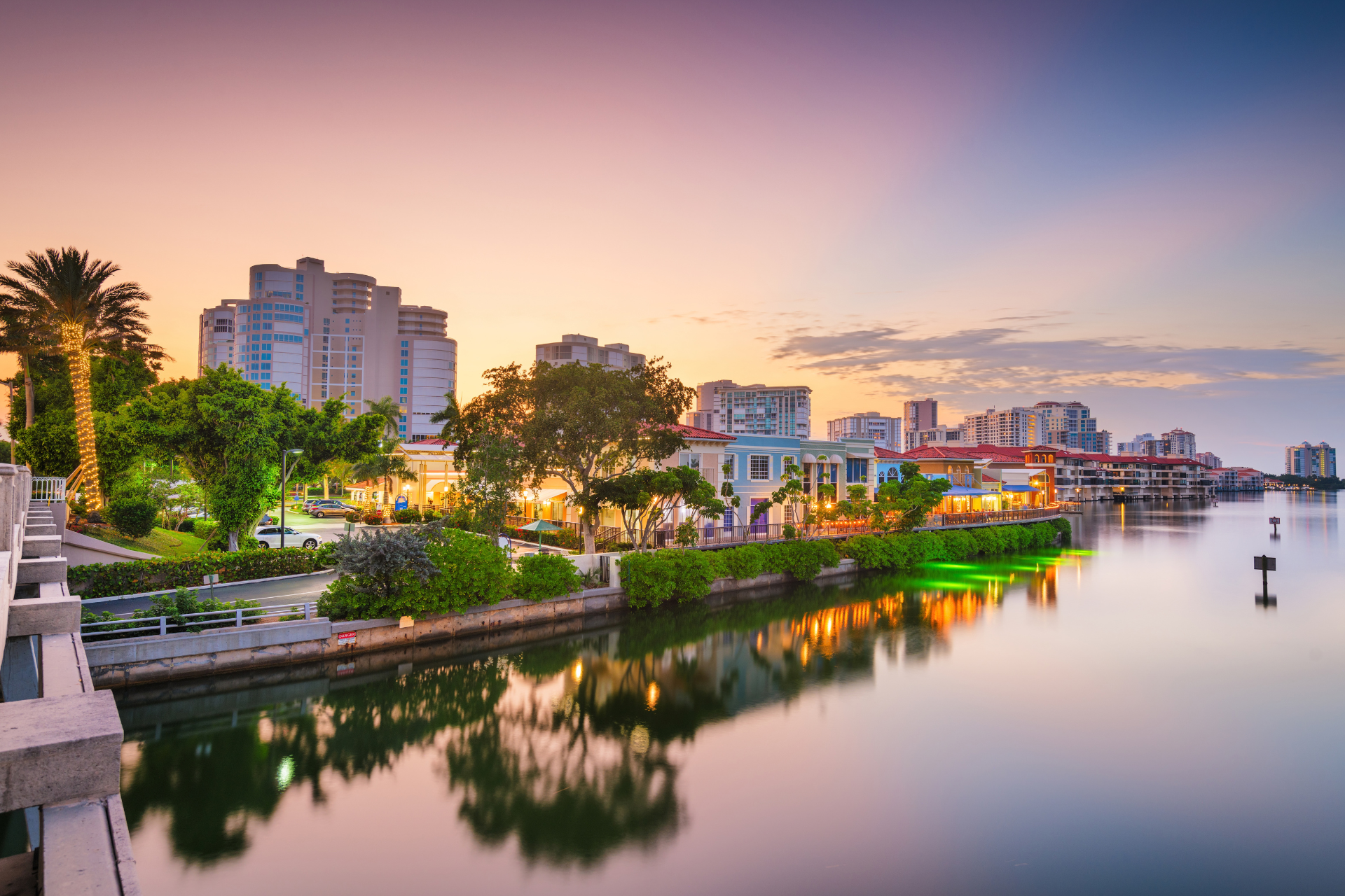 Waterfront residential and commercial district in Naples, Florida at sunset, with palm trees, luxury buildings, and reflections on calm water.