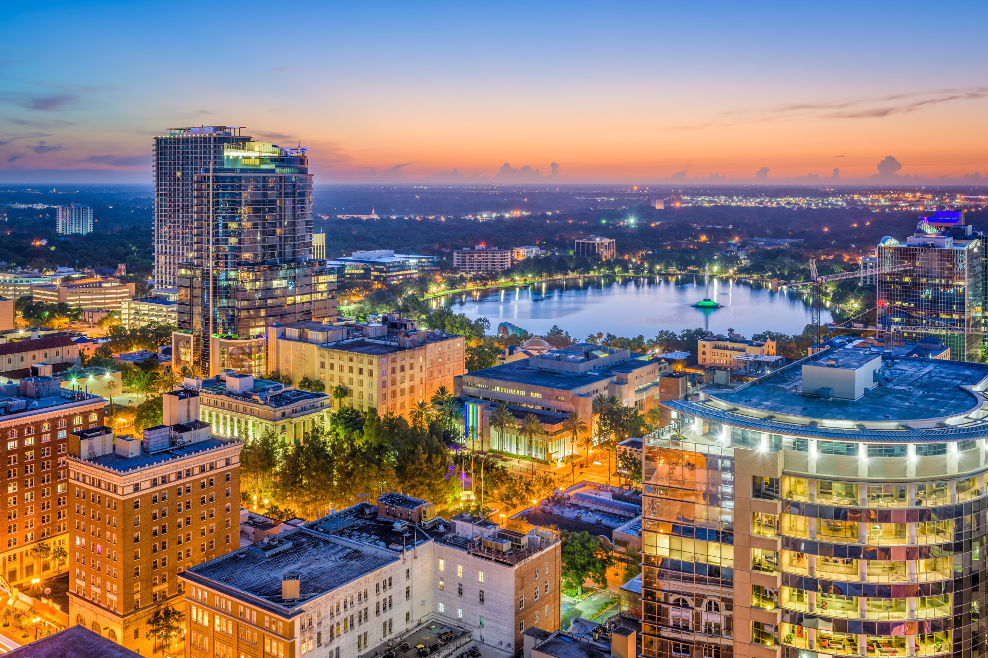 Evening aerial view of downtown Orlando, Florida, with illuminated high-rise buildings surrounding Lake Eola at dusk.