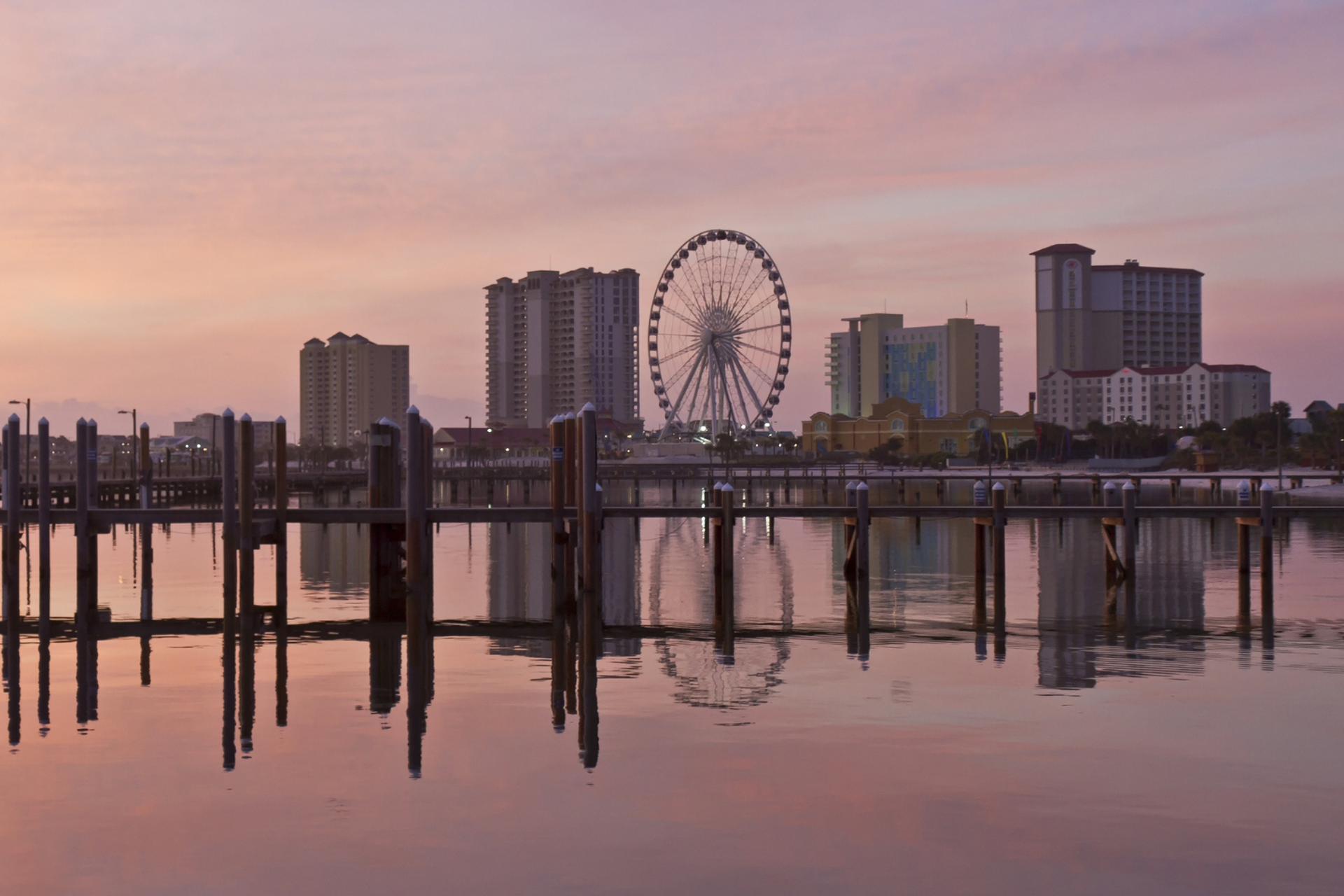 Pensacola, Florida waterfront at sunrise, with marina docks, a Ferris wheel, hotel buildings, and calm water reflections.