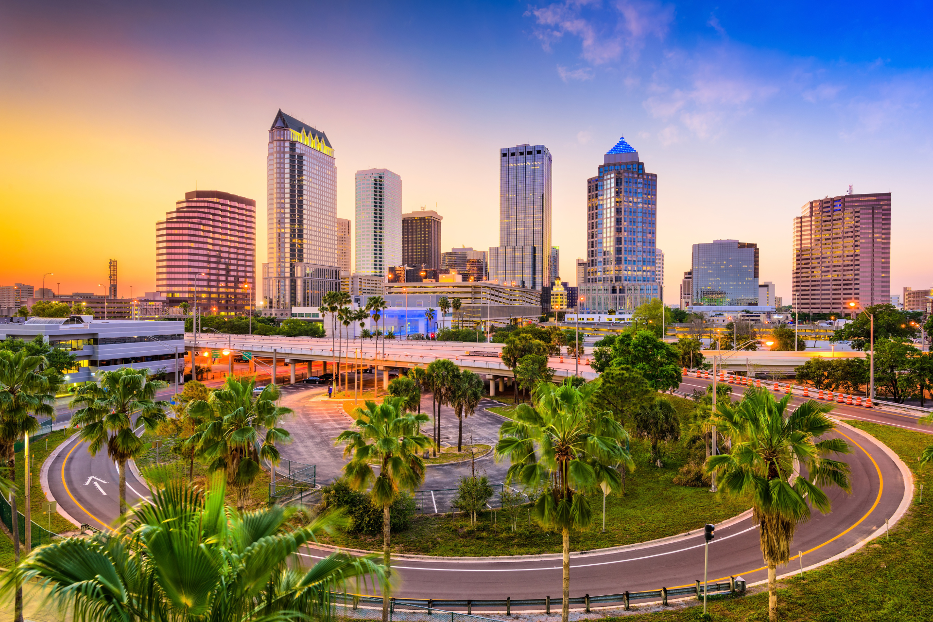 Downtown Tampa, Florida skyline at sunset, featuring modern office towers, palm trees, and elevated highways in an urban setting.
