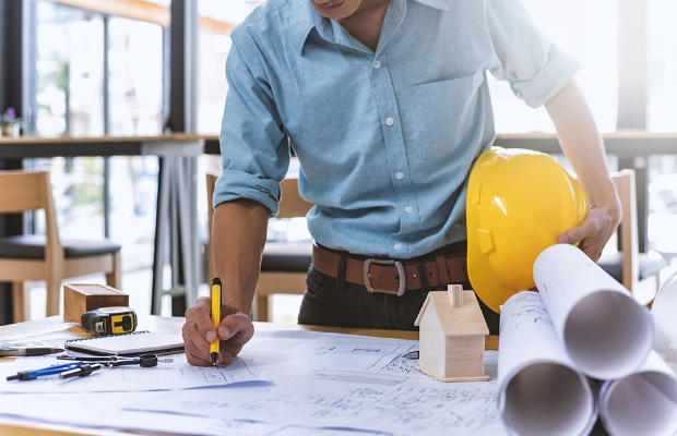 A contractor looking over architectural drawings while holding a hardhat in one hand and a pencil in the other. A contractor looking over architectural drawings while holding a hardhat in one hand and a pencil in the other.
