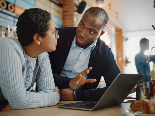 A man and a woman engaged in a discussion at a café, leaning over a table with a laptop open. A man and a woman engaged in a discussion at a café, leaning over a table with a laptop open.